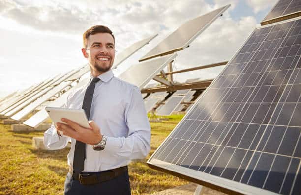 Positive male entrepreneur in formal wear browsing tablet while working in filed with modern photovoltaic panels and looking away